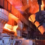 A crane-hung ladle beside a glowing converter inside a steel plant