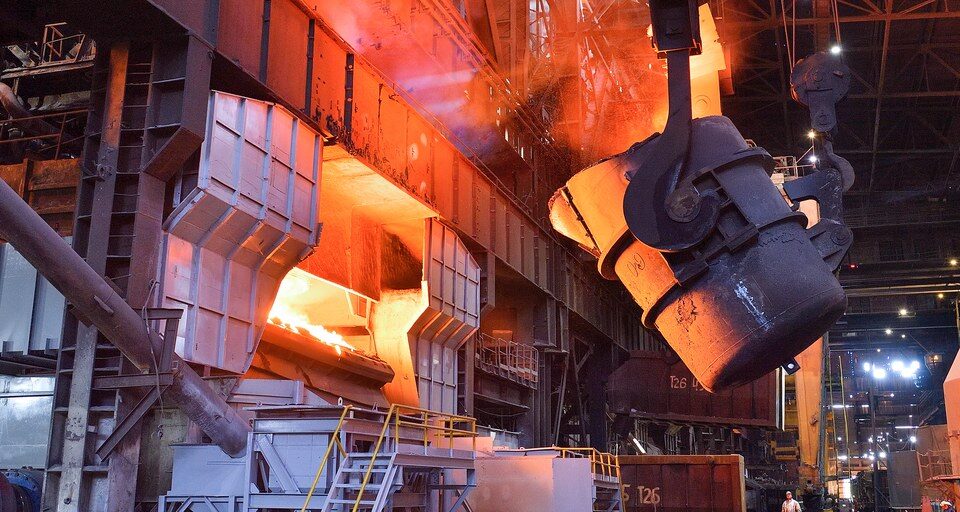 A crane-hung ladle beside a glowing converter inside a steel plant